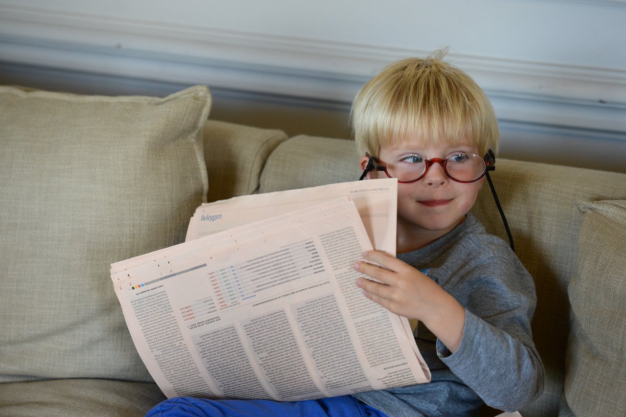 A young child with red glasses sits on a couch, smiling while holding and looking at a newspaper.