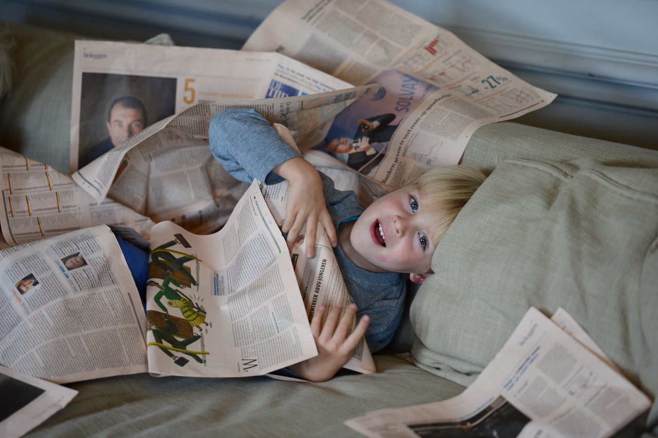 A smiling child lies on a couch, playfully covered in newspapers.