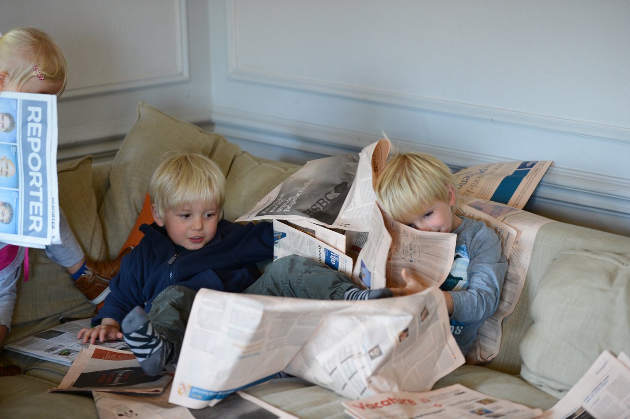 Two young children sit on a couch, playing with newspapers, while another child holds up a newspaper nearby.