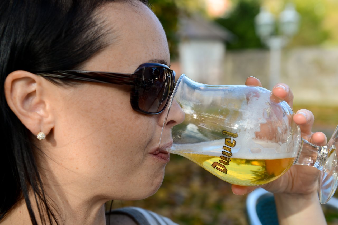 A woman wearing sunglasses drinks beer from a glass with "Duvel" written on it during an outdoor gathering.
