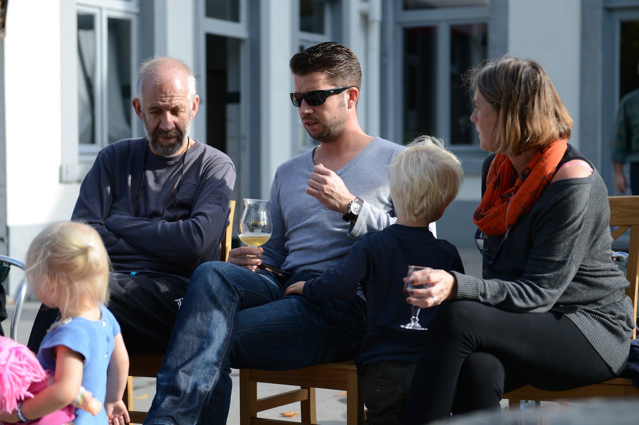 A group of adults and children sit outside, talking and holding drinks during a family gathering.