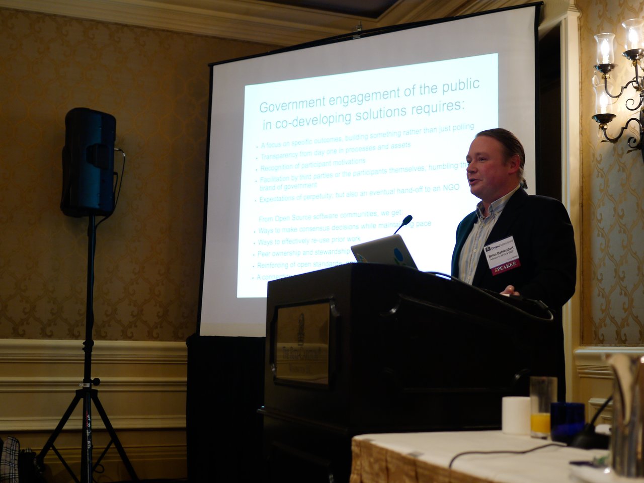 A speaker stands at a podium, presenting a slide about public engagement in co-developing solutions at a conference.