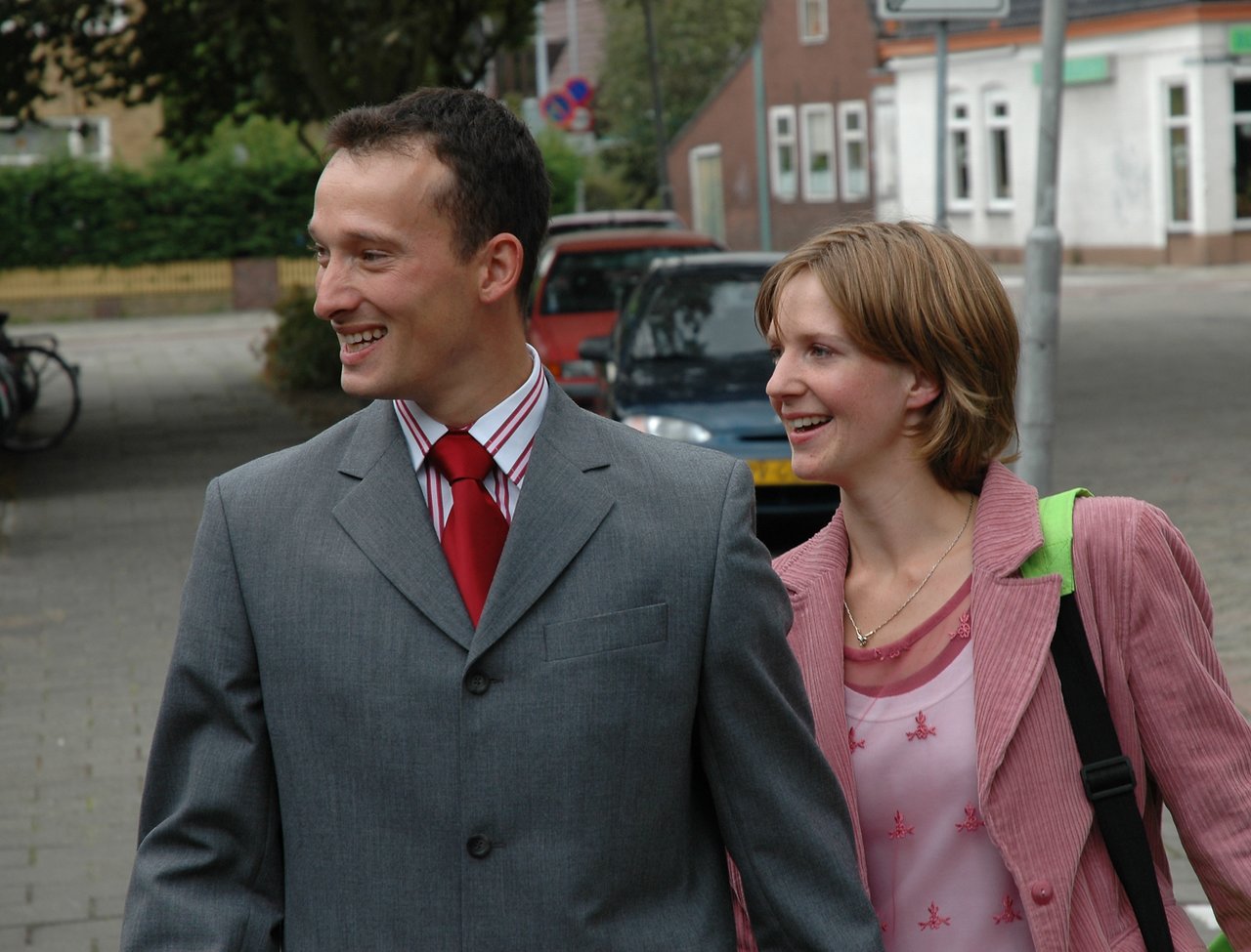 A man in a suit and a woman in a pink jacket walk together, smiling and looking to the side.
