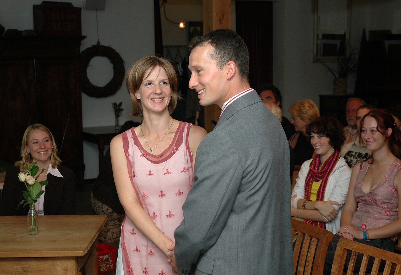 A smiling couple holds hands during a wedding ceremony, surrounded by seated guests in a warmly lit indoor setting.