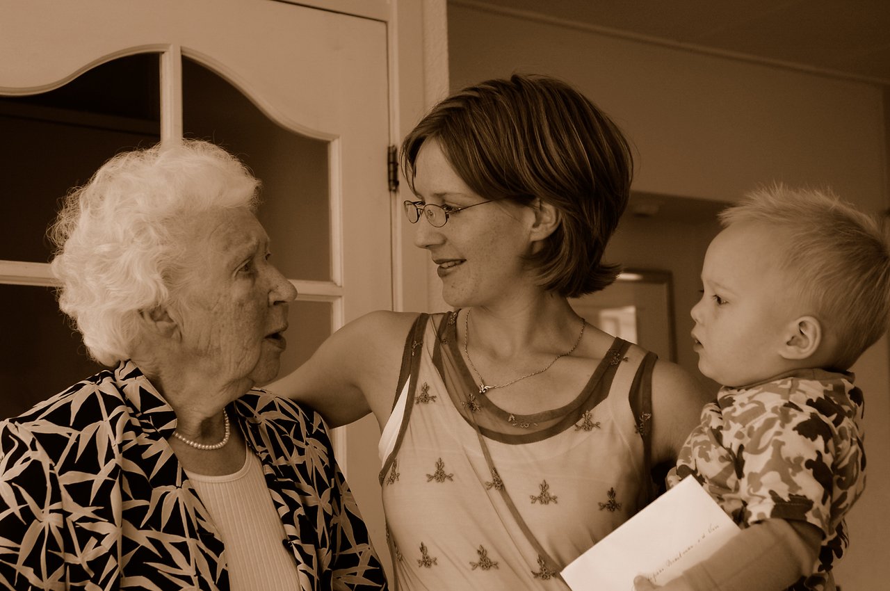 A woman holding a baby smiles while talking to an elderly woman indoors.