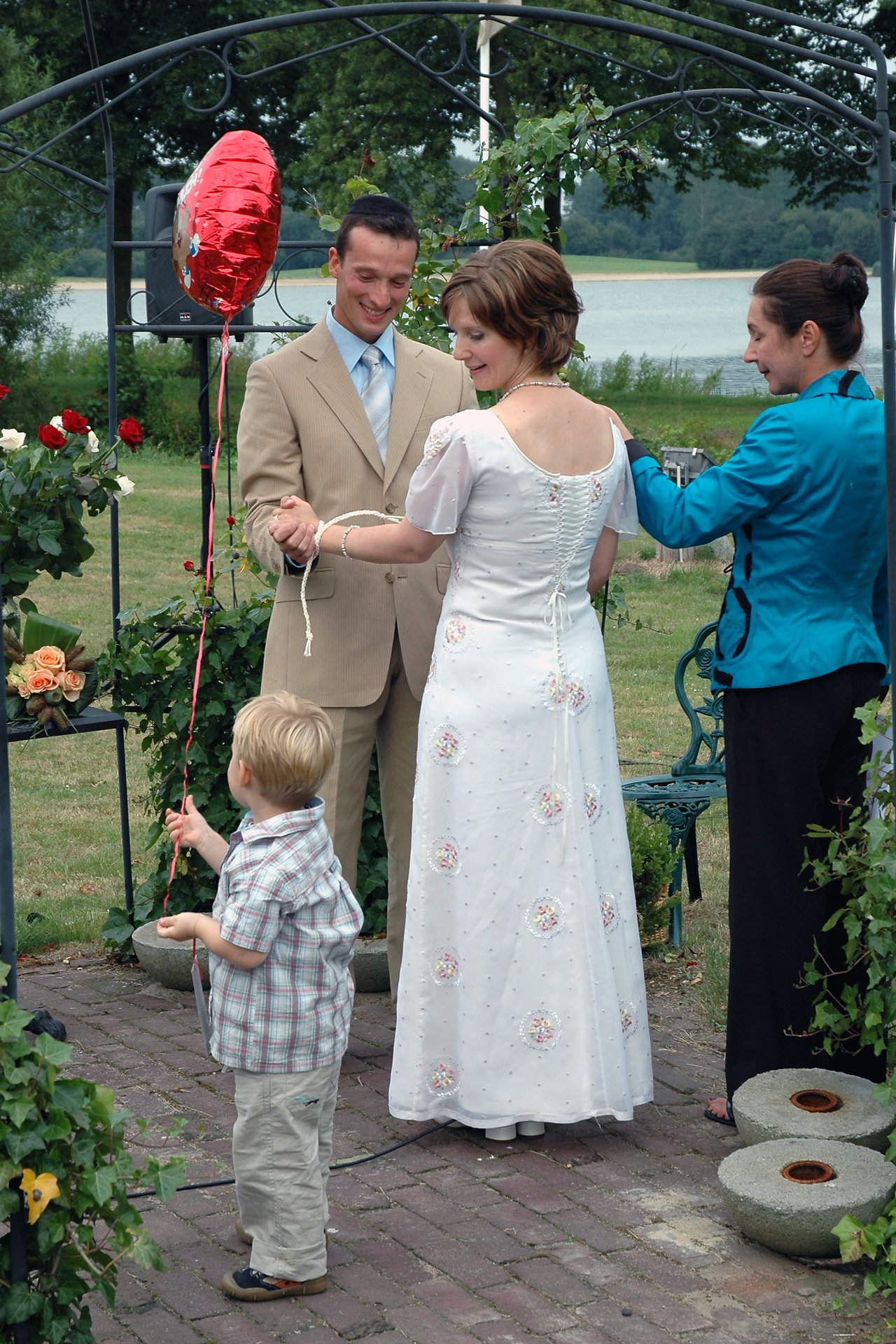 A bride and groom hold hands during their wedding ceremony while a woman assists and a child holds a balloon.