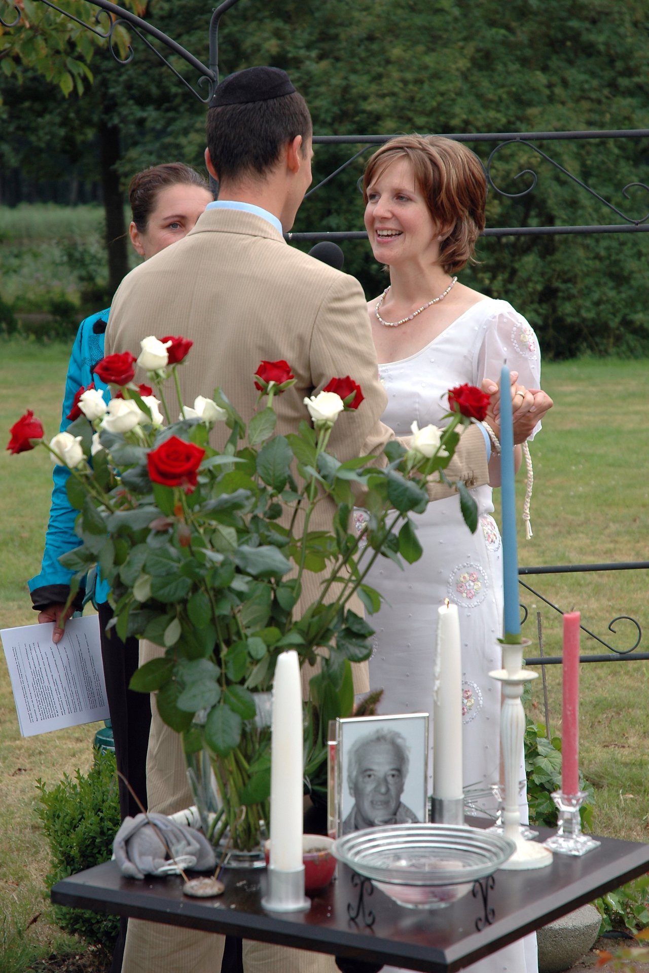A bride and groom stand facing each other, holding hands and smiling during their wedding ceremony outdoors.