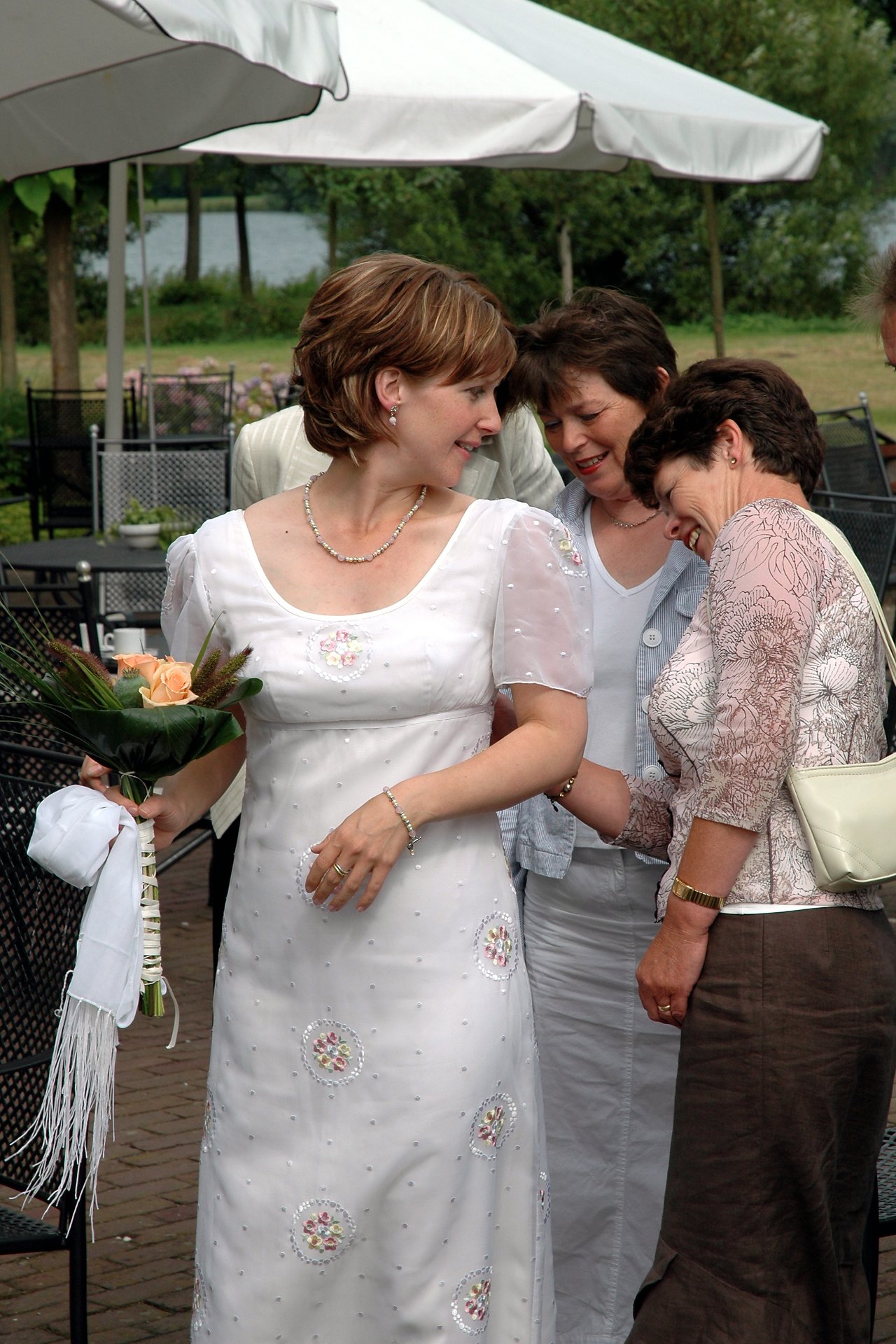 A bride in a white dress holds a bouquet while laughing with two women adjusting her gown.