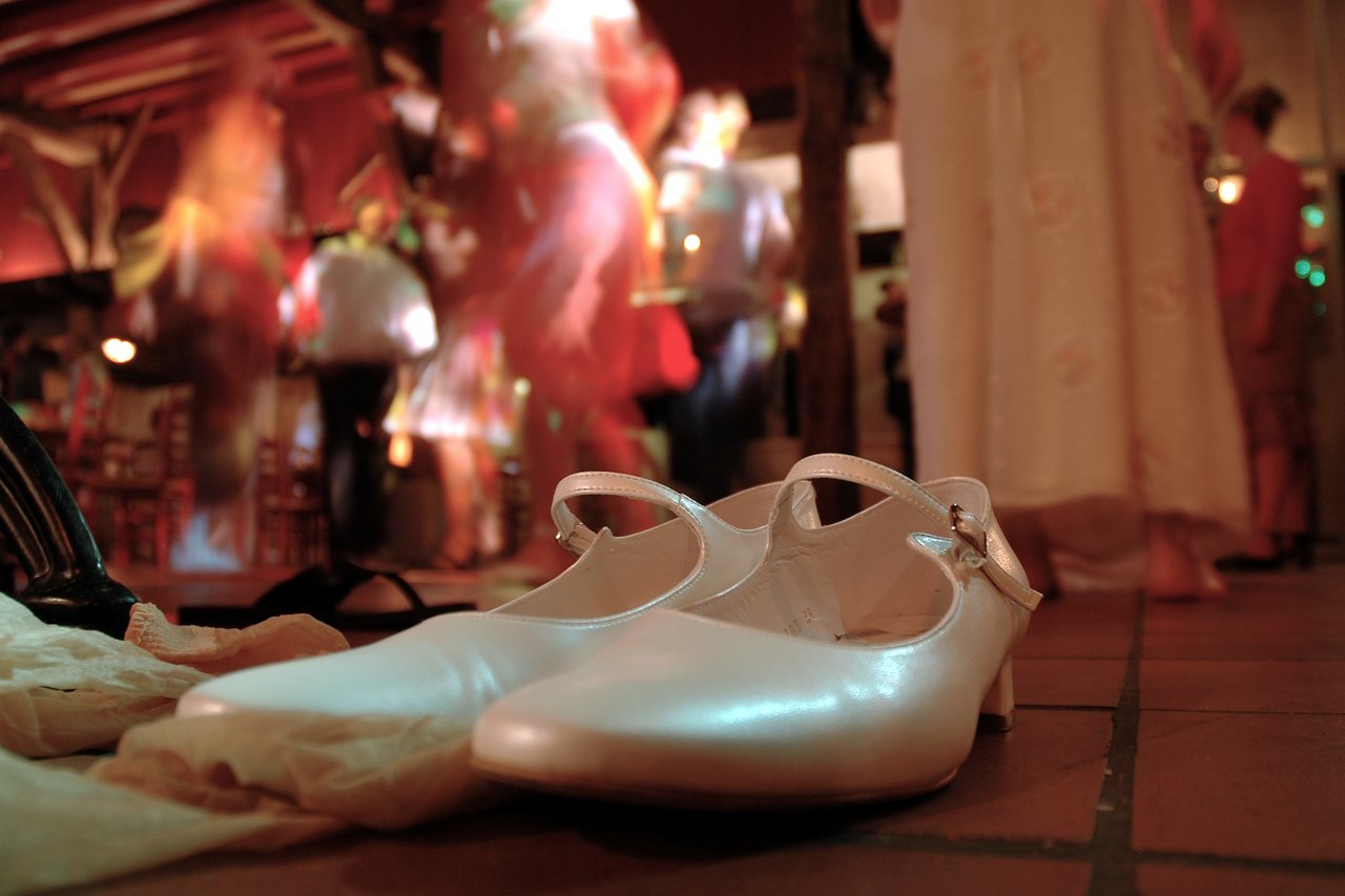 White wedding shoes rest on the floor as people dance in the background at a lively reception.