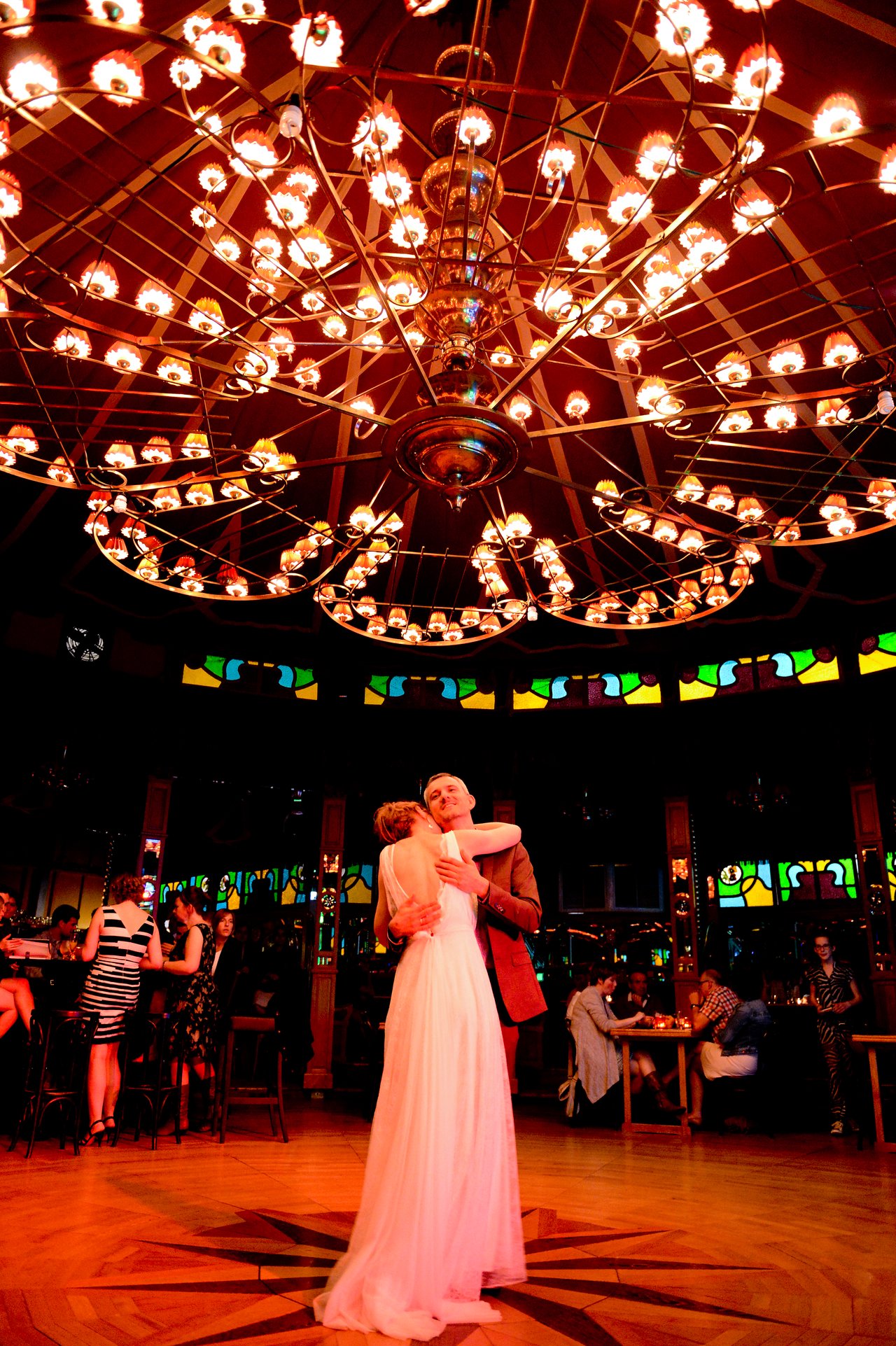 The bride and groom share a dance under a large chandelier in a warmly lit wedding venue.