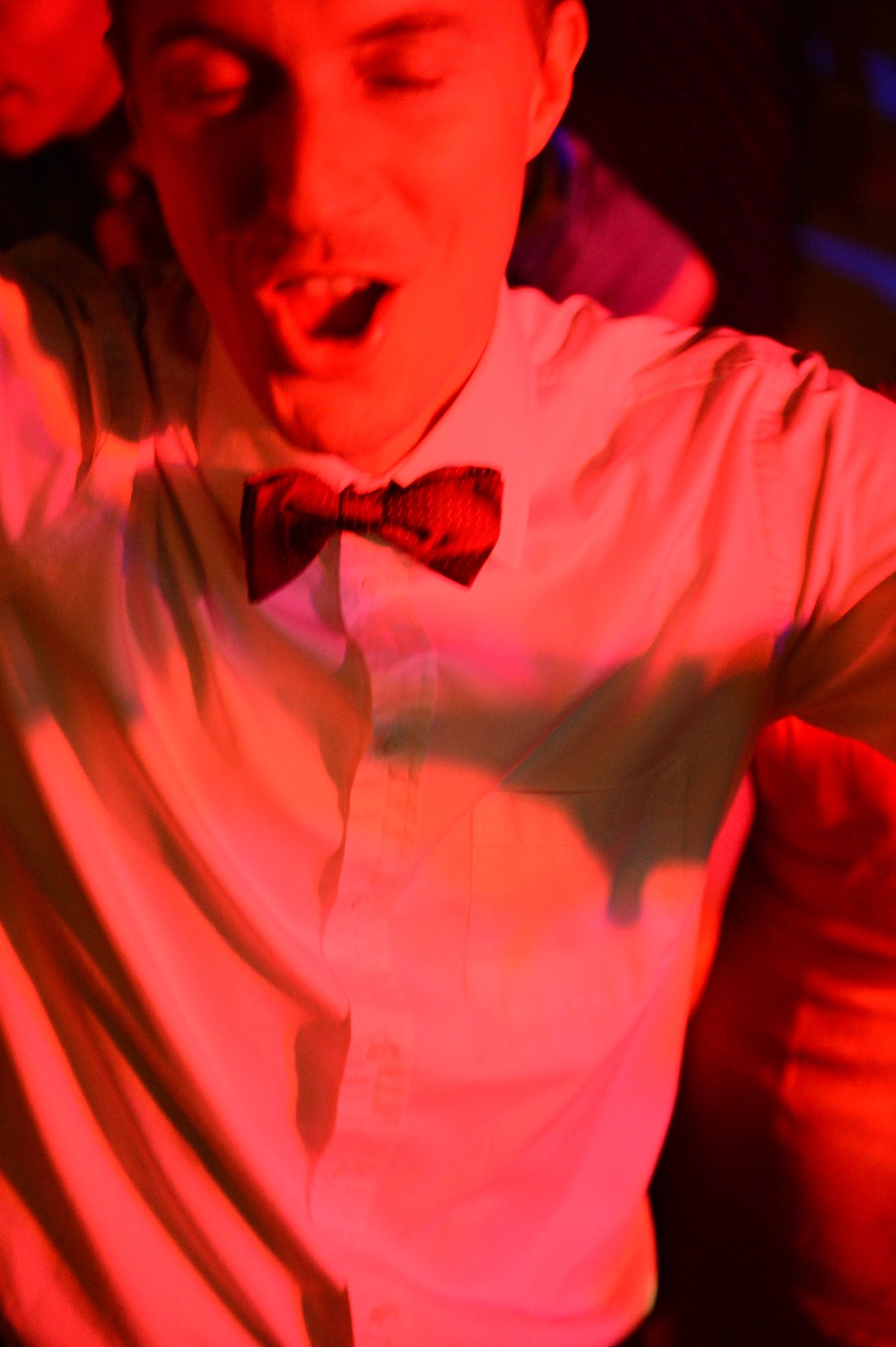 A man in a white shirt and bow tie dances with his mouth open under red lighting.