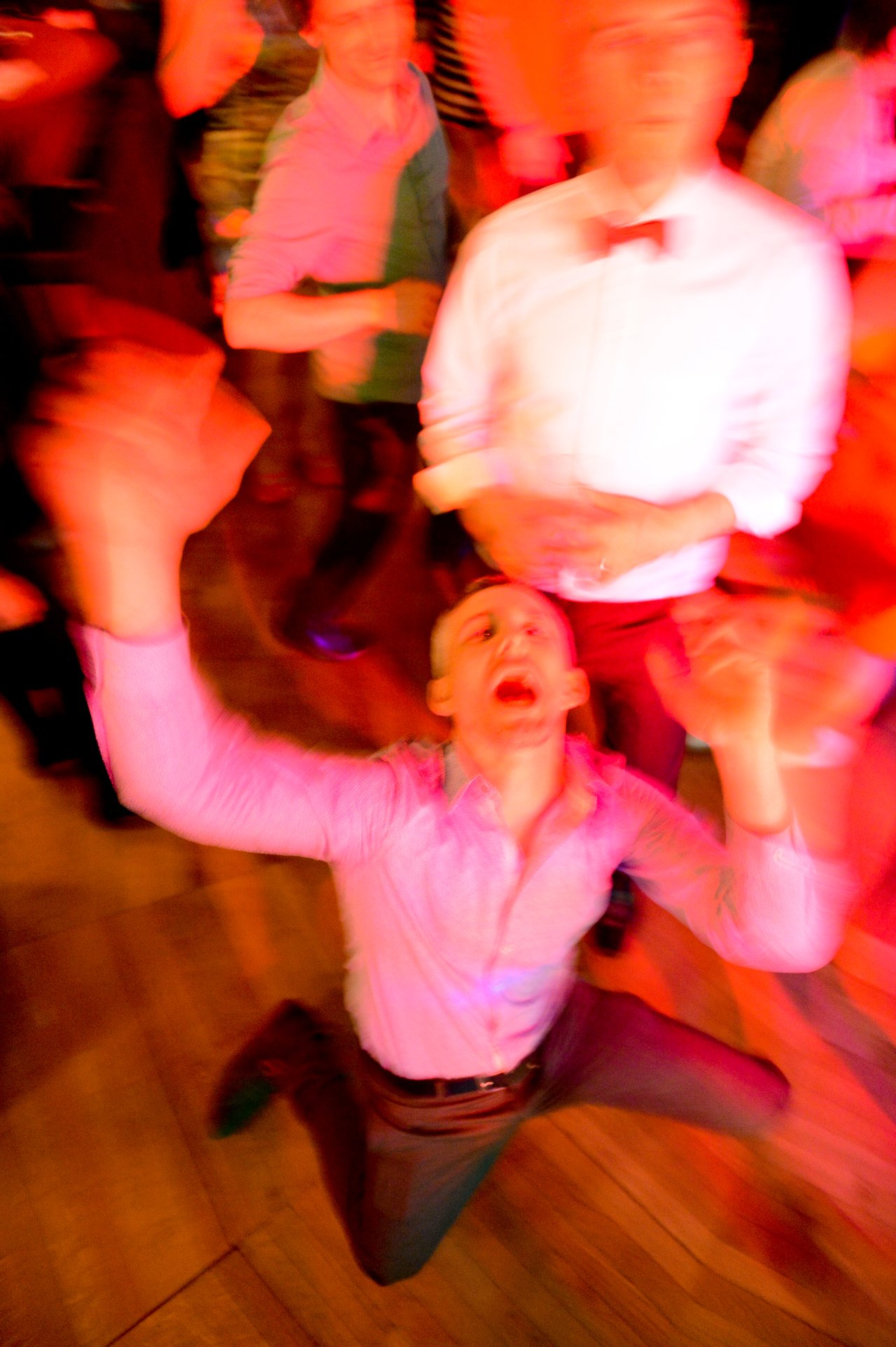 A man kneels on the dance floor with arms raised, surrounded by wedding guests in motion.