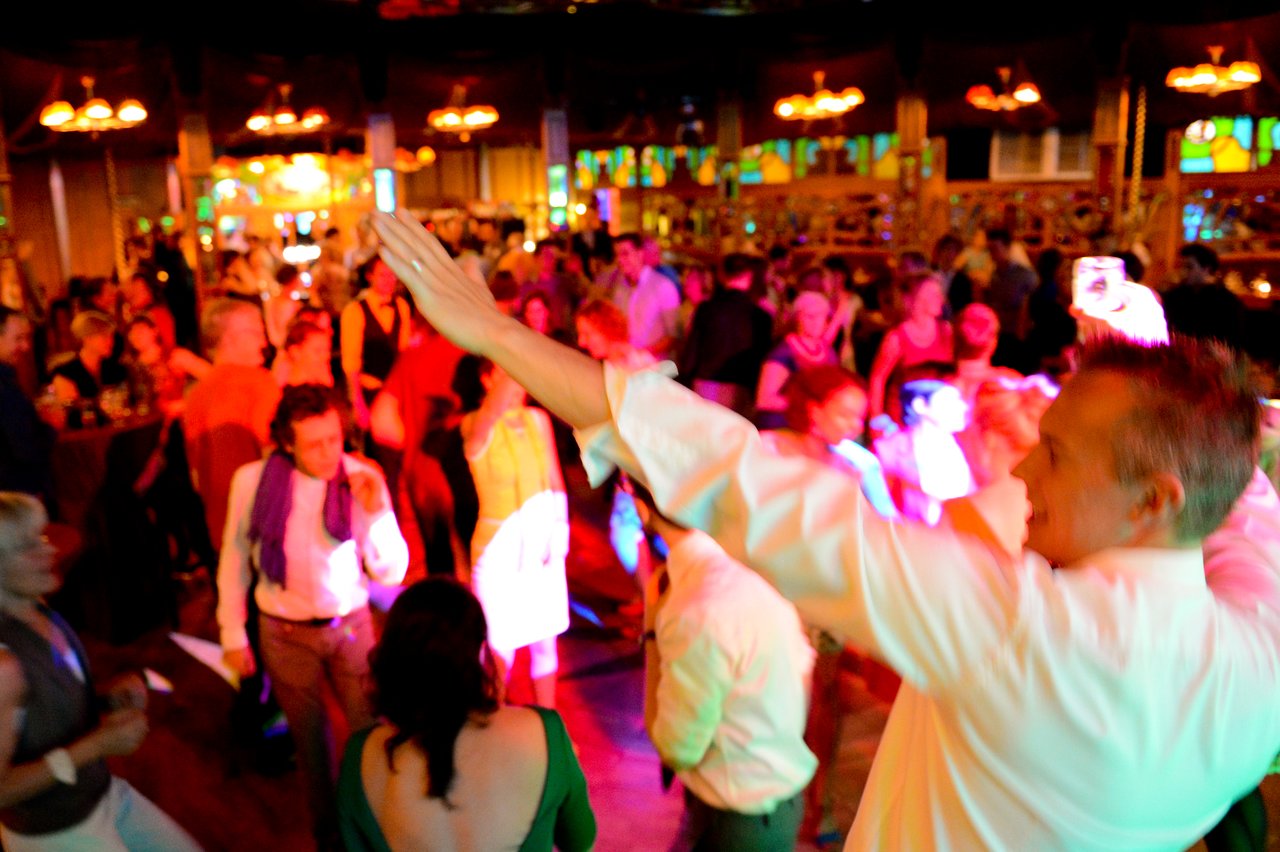 A man in a white shirt raises his hands while guests dance at a lively wedding reception.