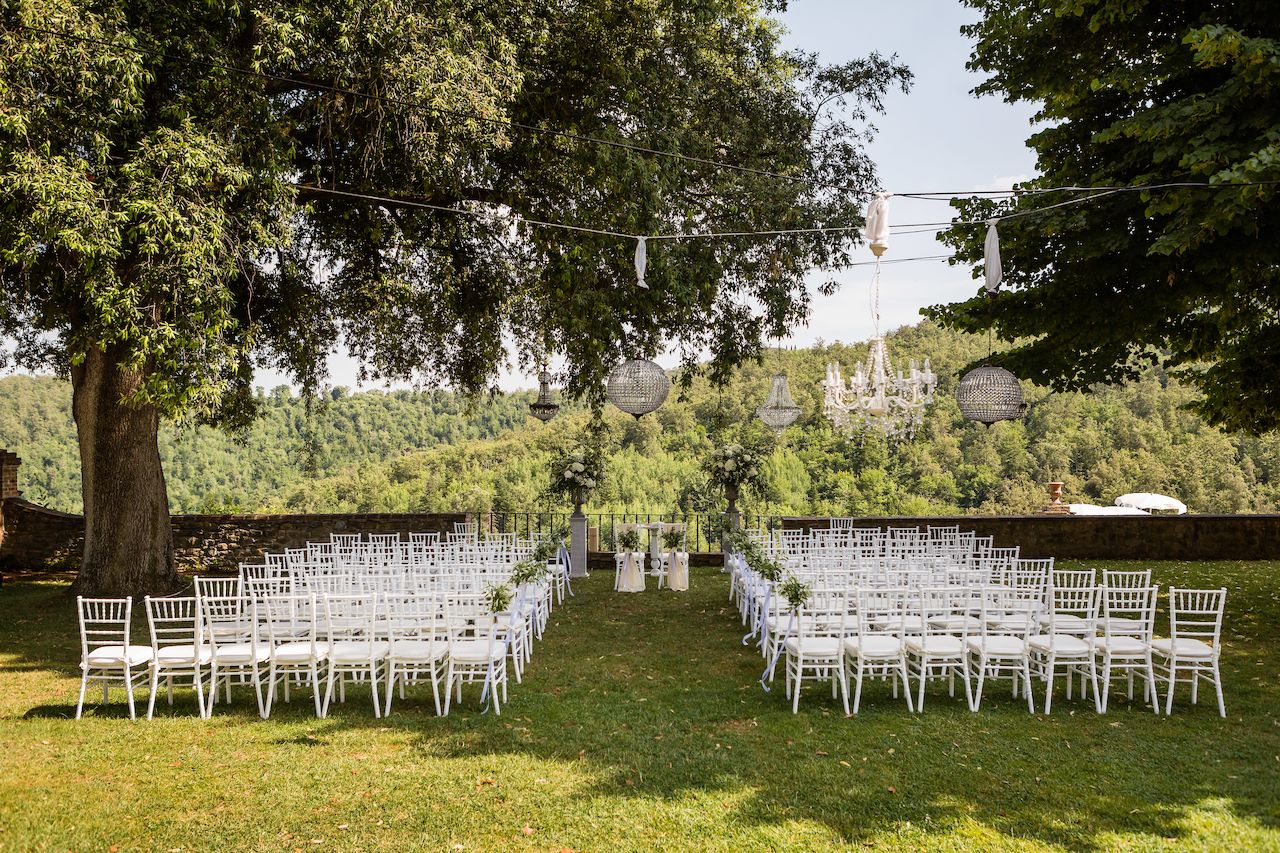 Outdoor wedding ceremony setup with white chairs arranged in rows, a decorated altar, and hanging chandeliers under trees.