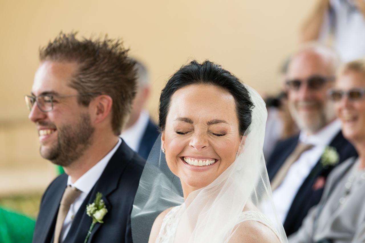 The bride smiles with her eyes closed, while the groom sits beside her, both dressed for the wedding ceremony.