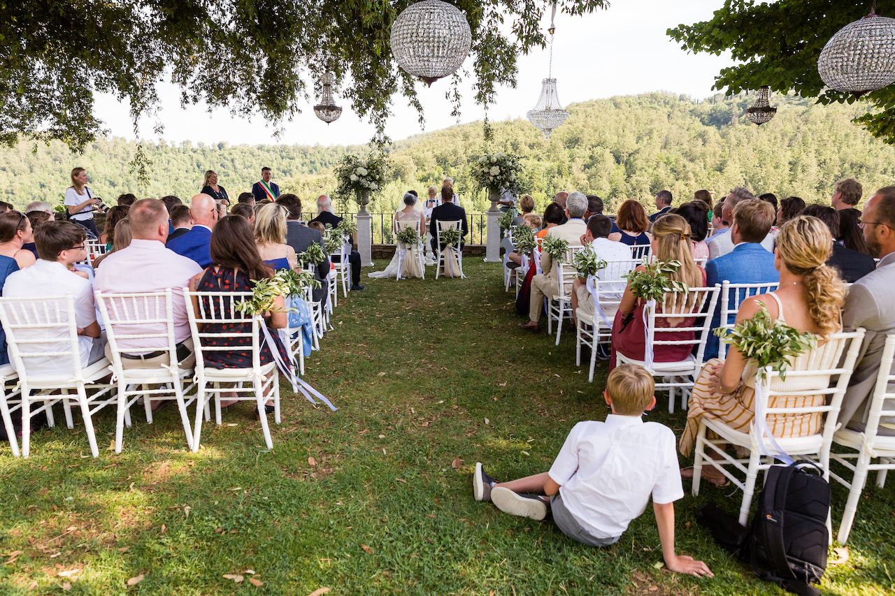 Wedding ceremony taking place outdoors with guests seated, facing the couple and officiant at the front.