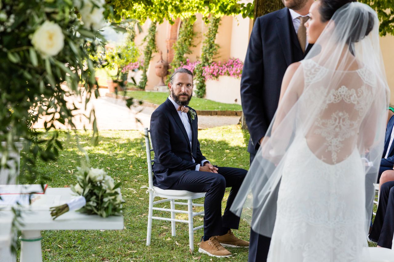 The groom sits on a chair, looking towards the bride, who stands in a white wedding dress with a veil.