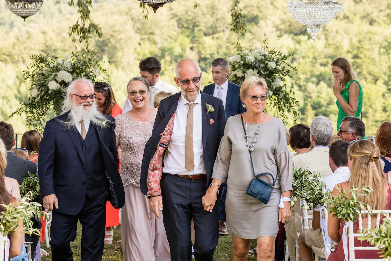 Four elegantly dressed guests walk down the aisle, smiling, at an outdoor wedding ceremony with seated attendees.