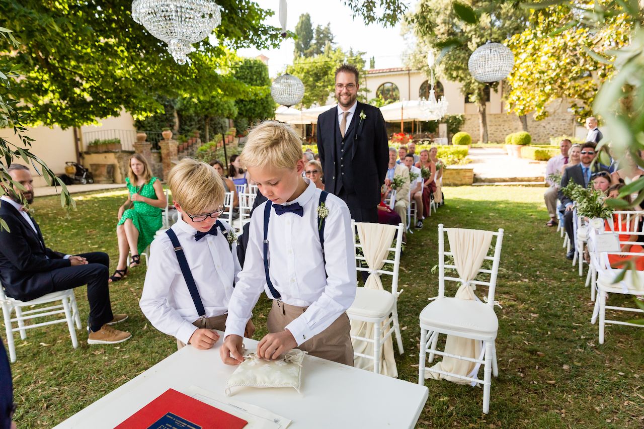 Two young boys in suspenders handle a ring pillow at an outdoor wedding ceremony, with guests watching.