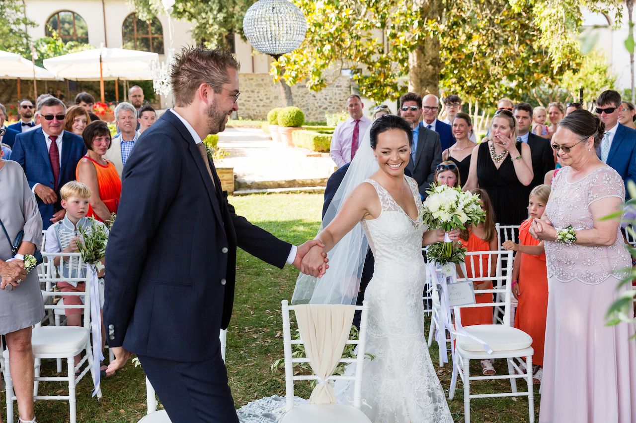 The groom walks toward the bride, holding her hand, as guests watch and smile during the wedding ceremony.