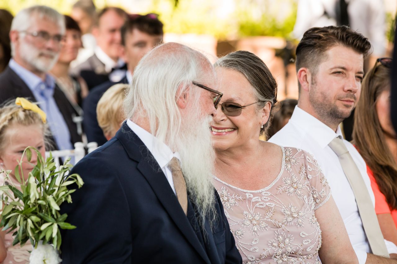 An older couple smiles at each other while seated at a wedding ceremony, surrounded by other guests.