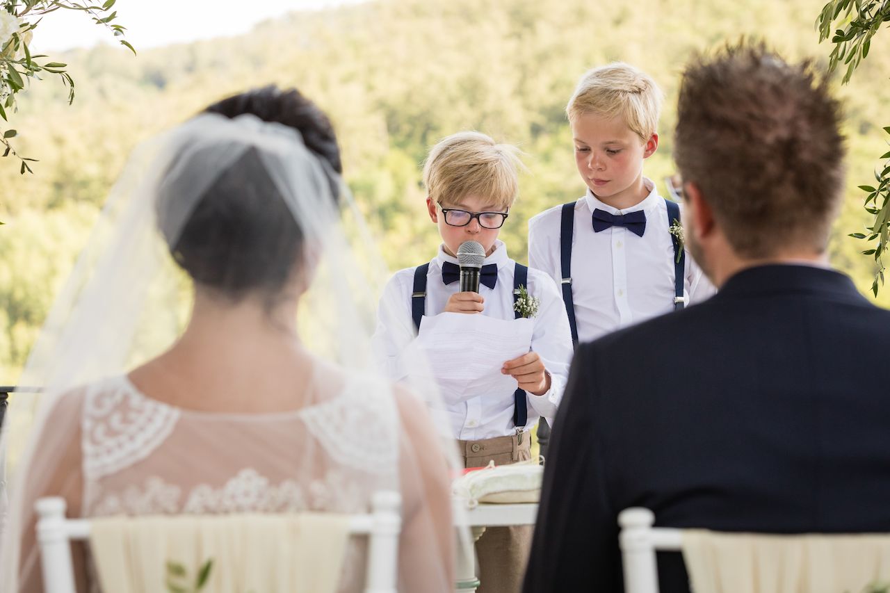 Two young boys in formal attire read a speech into a microphone during a wedding ceremony.