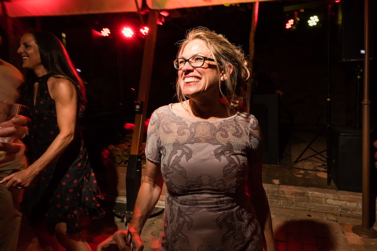 A woman in glasses and a patterned dress smiles while dancing at a wedding reception with others.