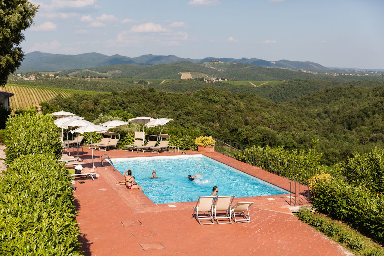 A few people relax in and around a swimming pool with a scenic hillside view in the background.