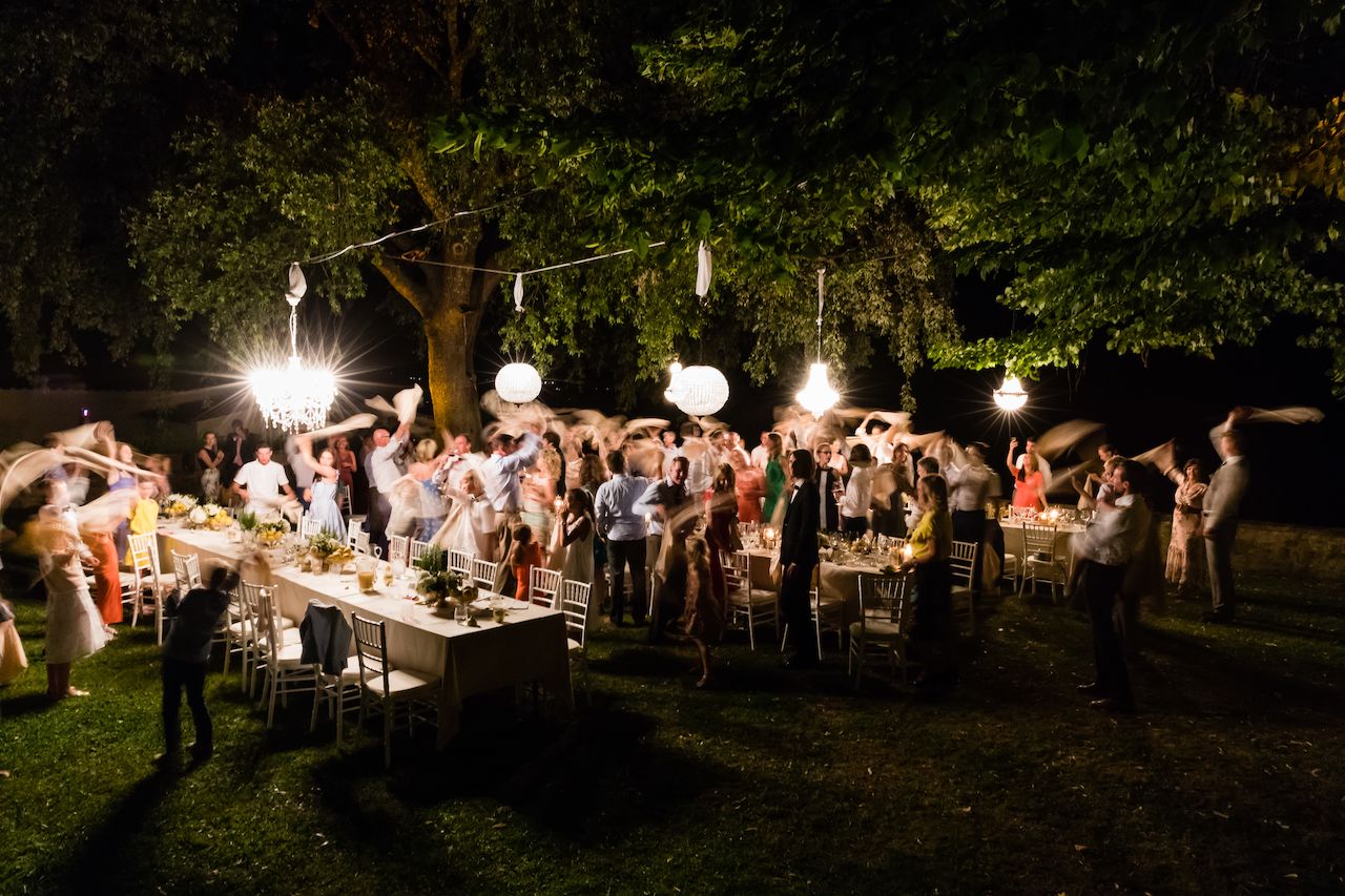 Wedding guests stand and wave napkins in the air during an outdoor dinner celebration under hanging lights.
