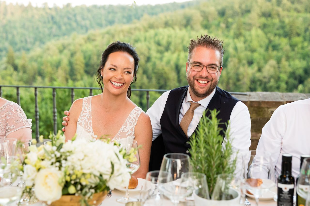 A smiling bride and groom sit together at a wedding dinner table, surrounded by flowers, drinks, and guests.
