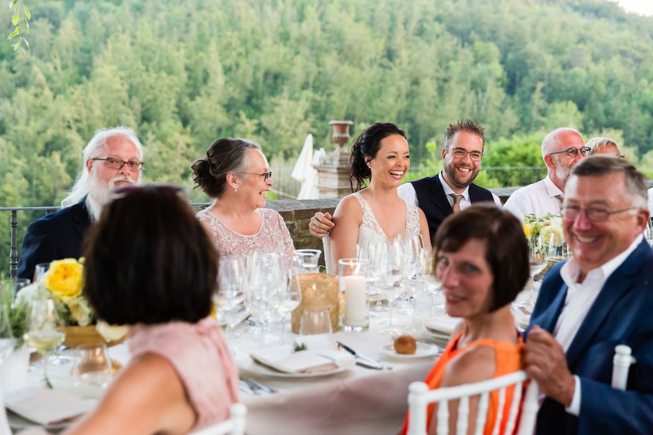 The bride and groom smile while seated at a dinner table with family and friends during their wedding celebration.