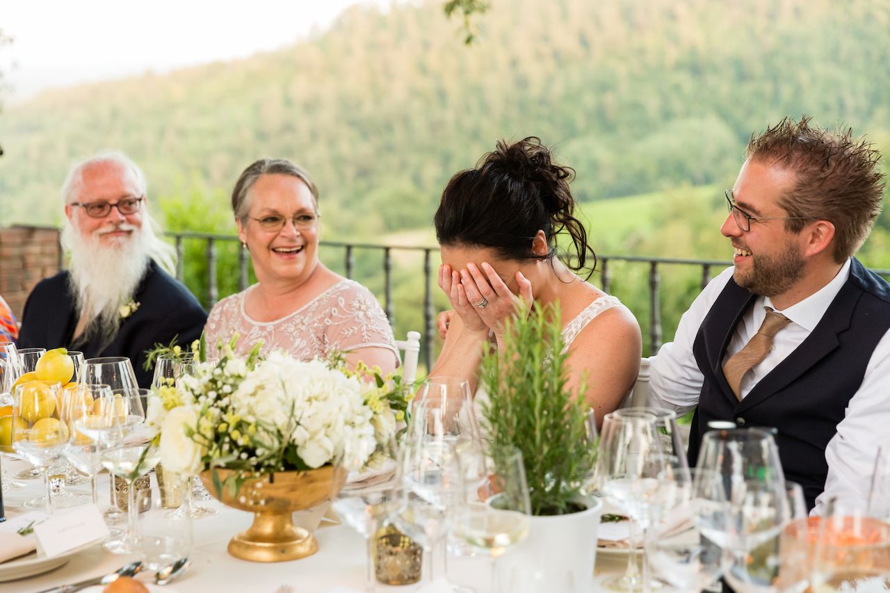 The bride covers her face while laughing at the dinner table, surrounded by smiling guests.