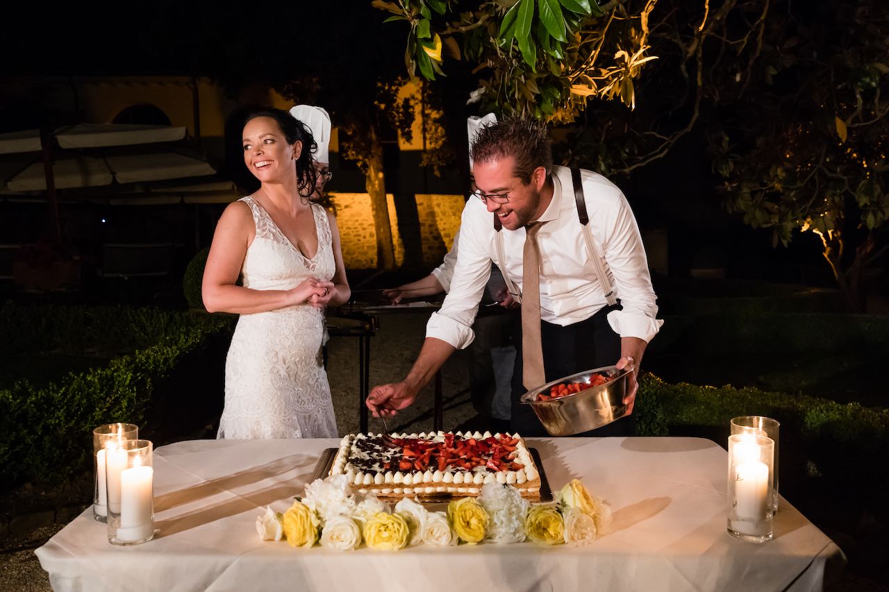 The bride smiles while the groom adds strawberries to a wedding cake at an outdoor dinner reception.