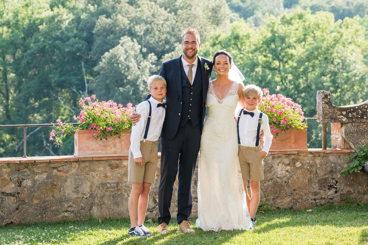 Bride and groom stand smiling with two young boys in suspenders, posing together for a wedding photo.