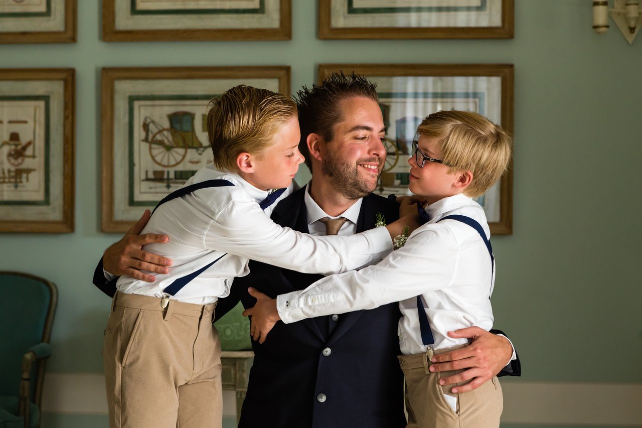 A man in a suit smiles while hugging two young boys dressed in suspenders and bow ties.
