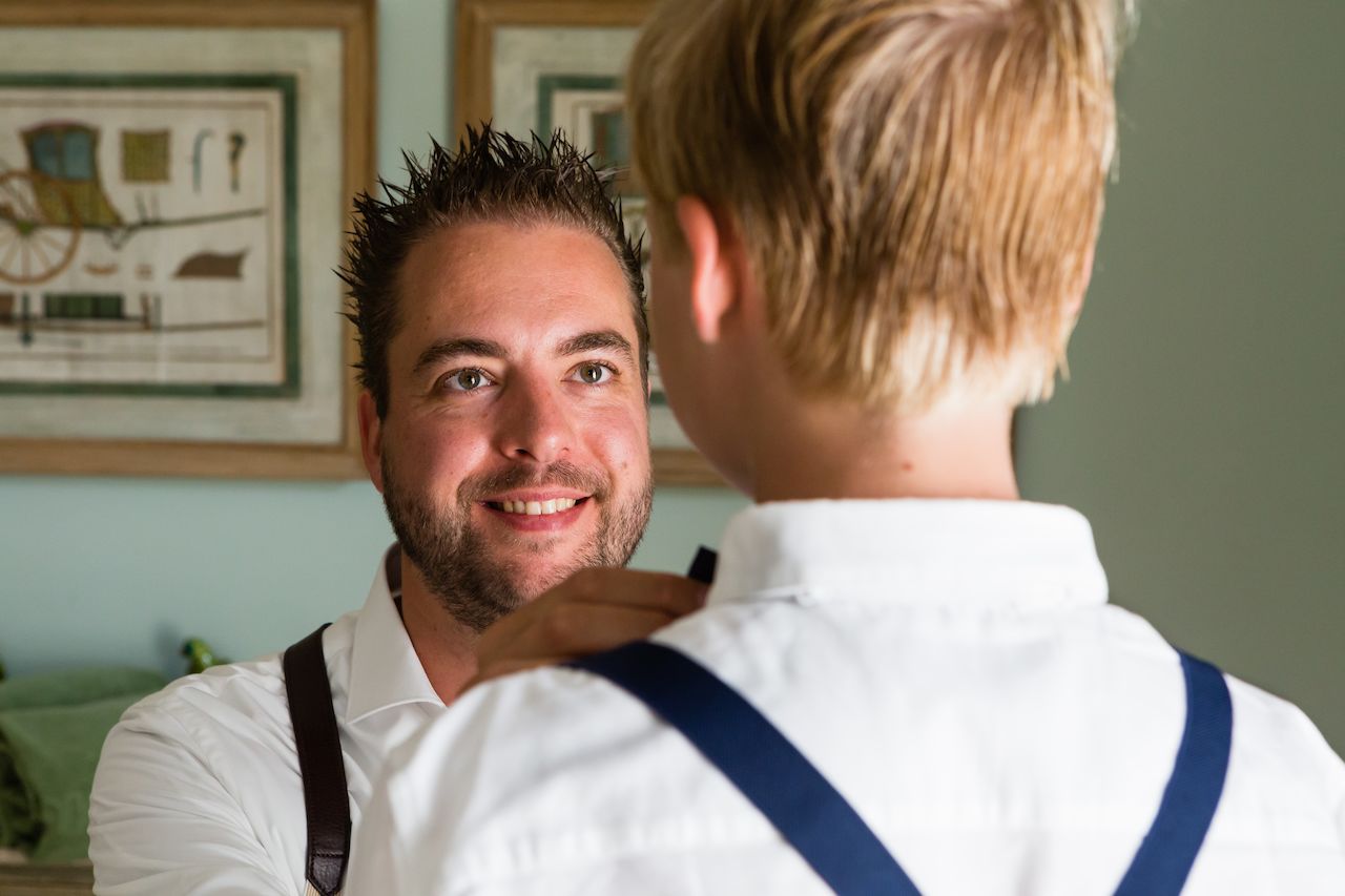 A man smiles while helping a young boy adjust his suspenders before the wedding.