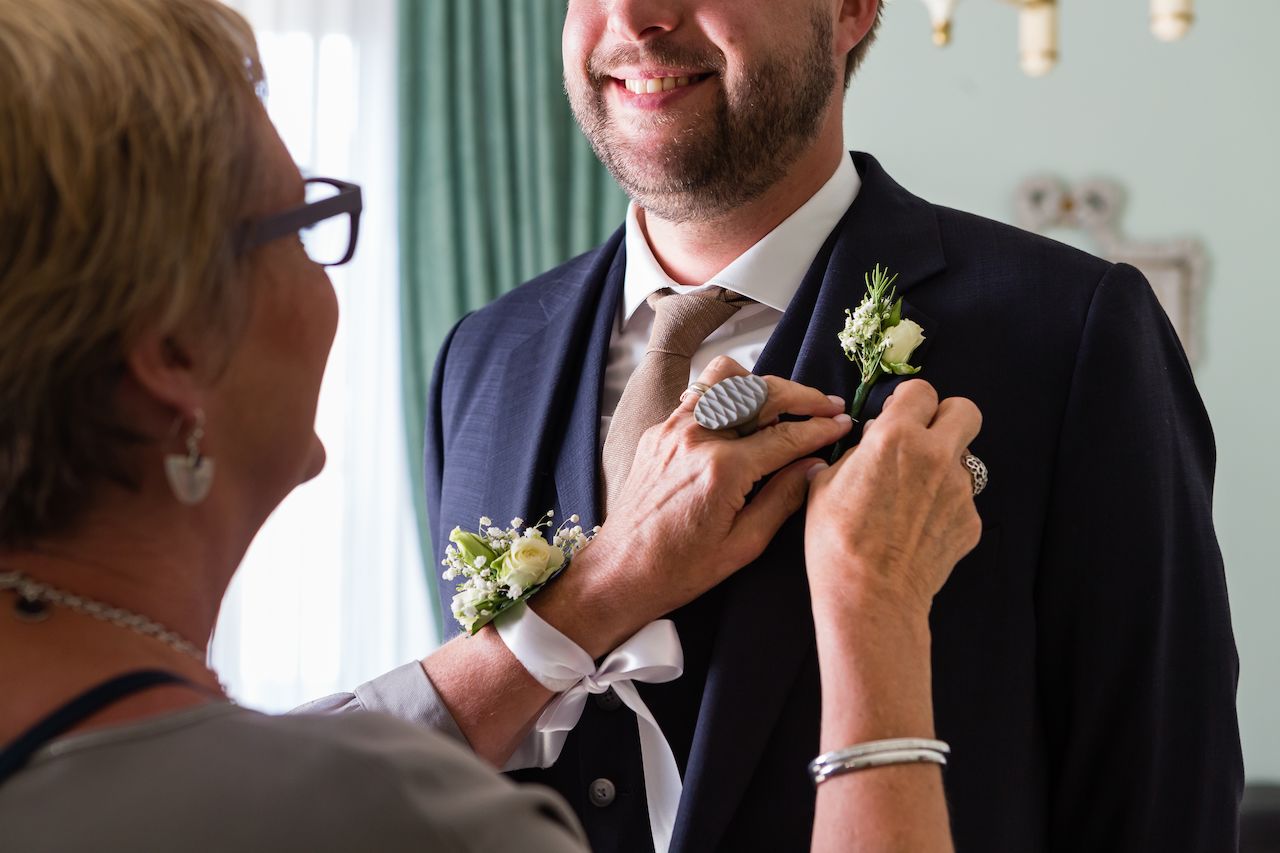 A woman pins a boutonniere on a smiling groom's suit jacket as they prepare for the wedding.