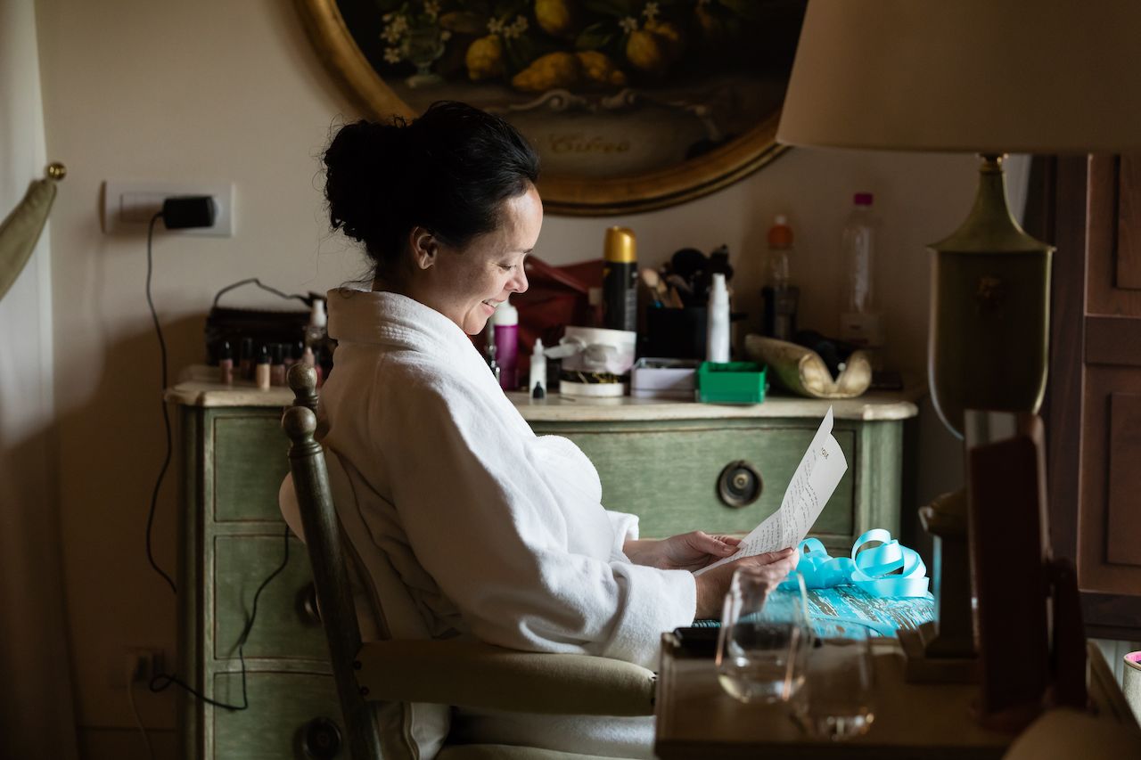 A woman in a robe sits at a vanity, smiling while reading a letter with a gift beside her.