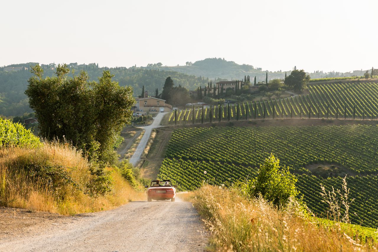 A couple drives a red convertible down a dirt road through a vineyard, leaving a trail of dust behind.