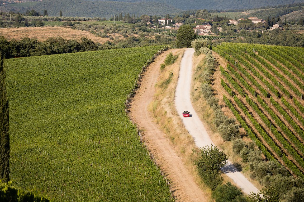A red convertible drives along a dirt road through a vineyard, with two people inside.