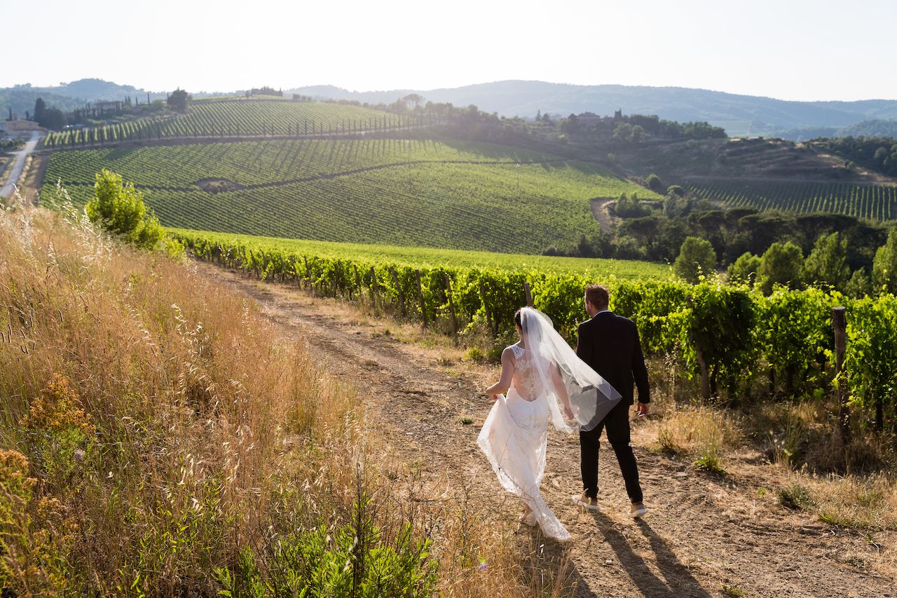 A bride and groom walk hand in hand along a dirt path through a vineyard.