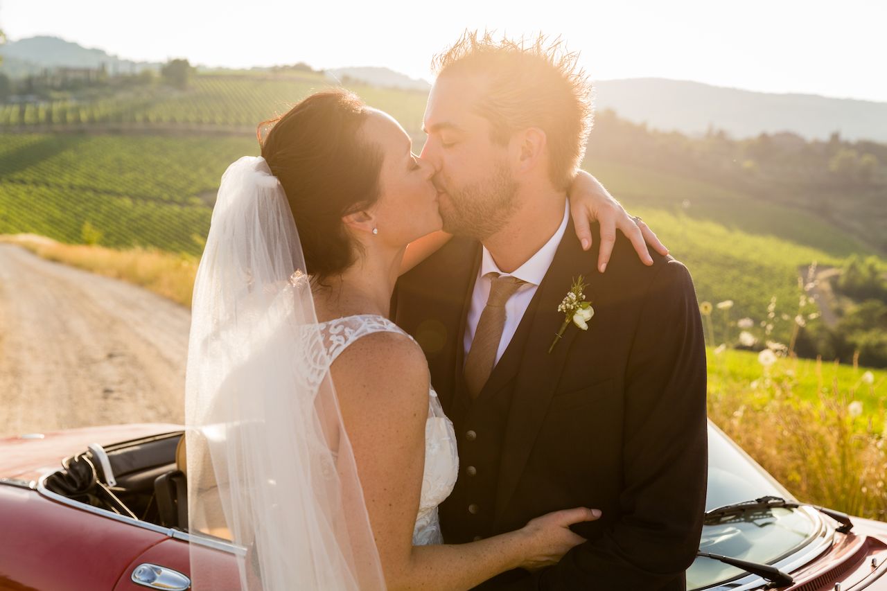 A bride and groom share a kiss in a vineyard, standing beside a red convertible car.