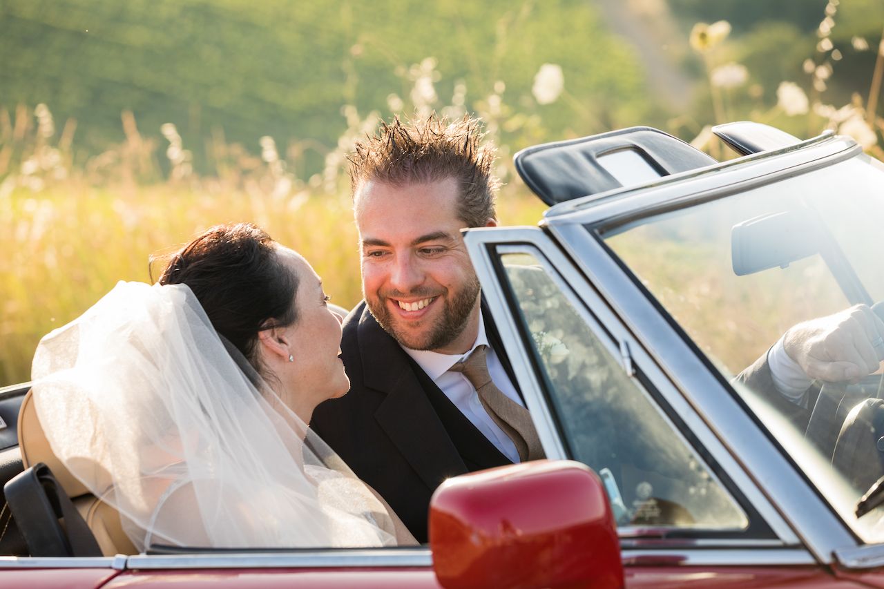 A bride and groom smile at each other while sitting in a red convertible with the top down.