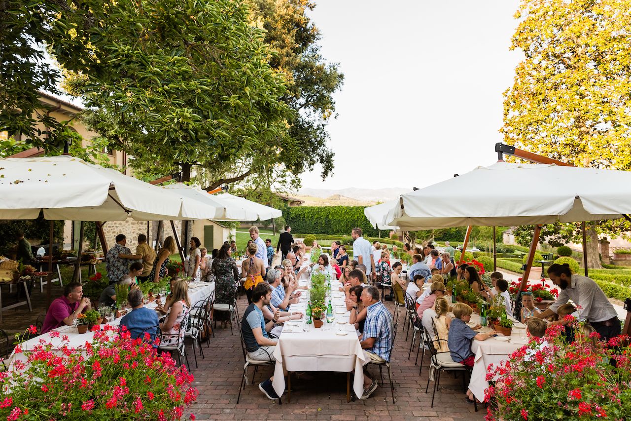 Guests sit at long tables under umbrellas, enjoying food and conversation at an outdoor wedding welcome barbecue.