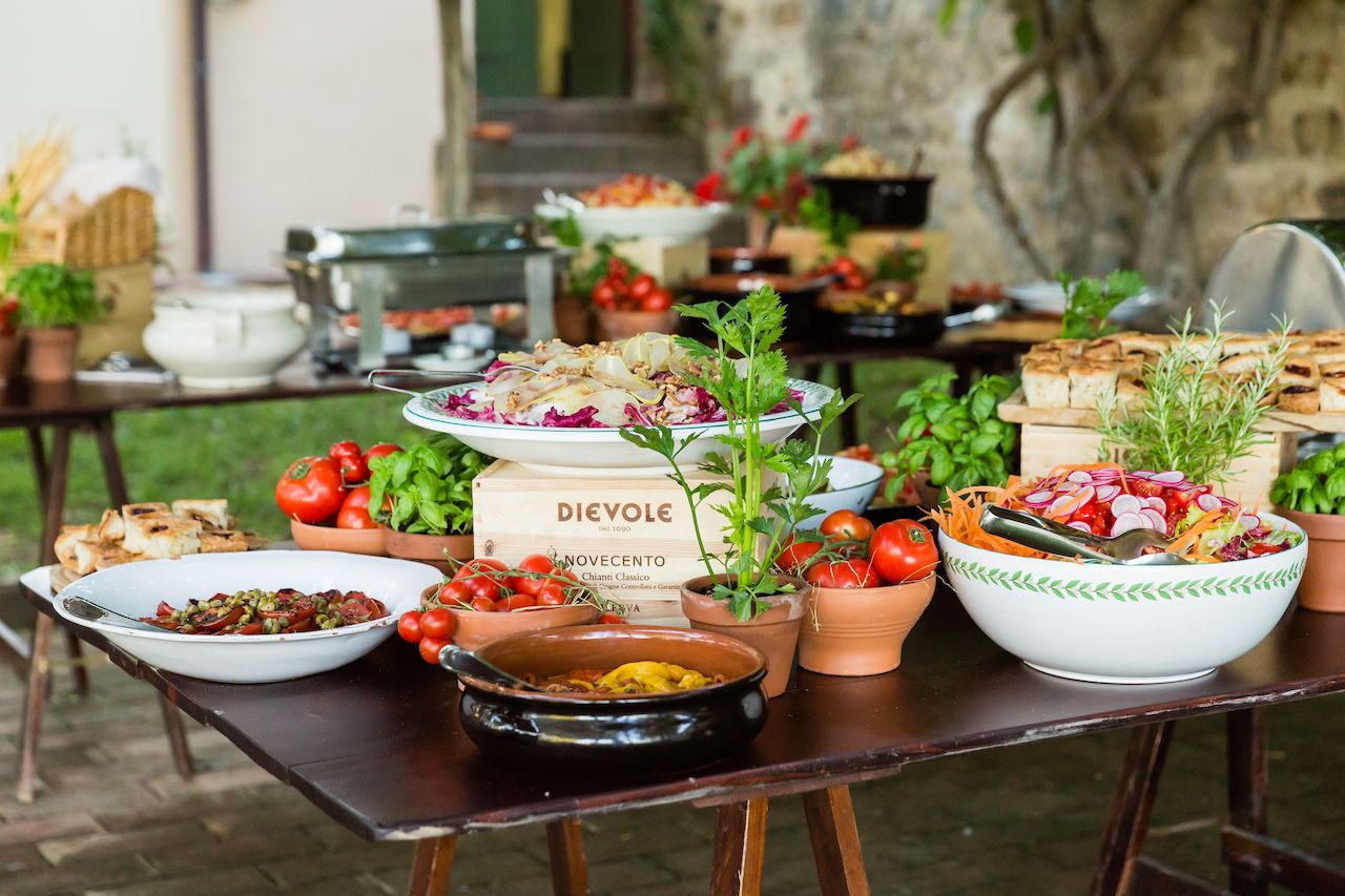 A buffet table with fresh salads, vegetables, and bread set up for an outdoor wedding welcome barbecue.