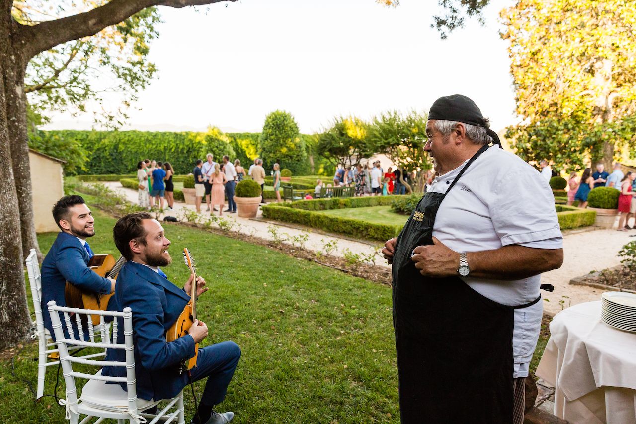 A chef in an apron talks to two musicians playing guitars, while guests mingle in the background.