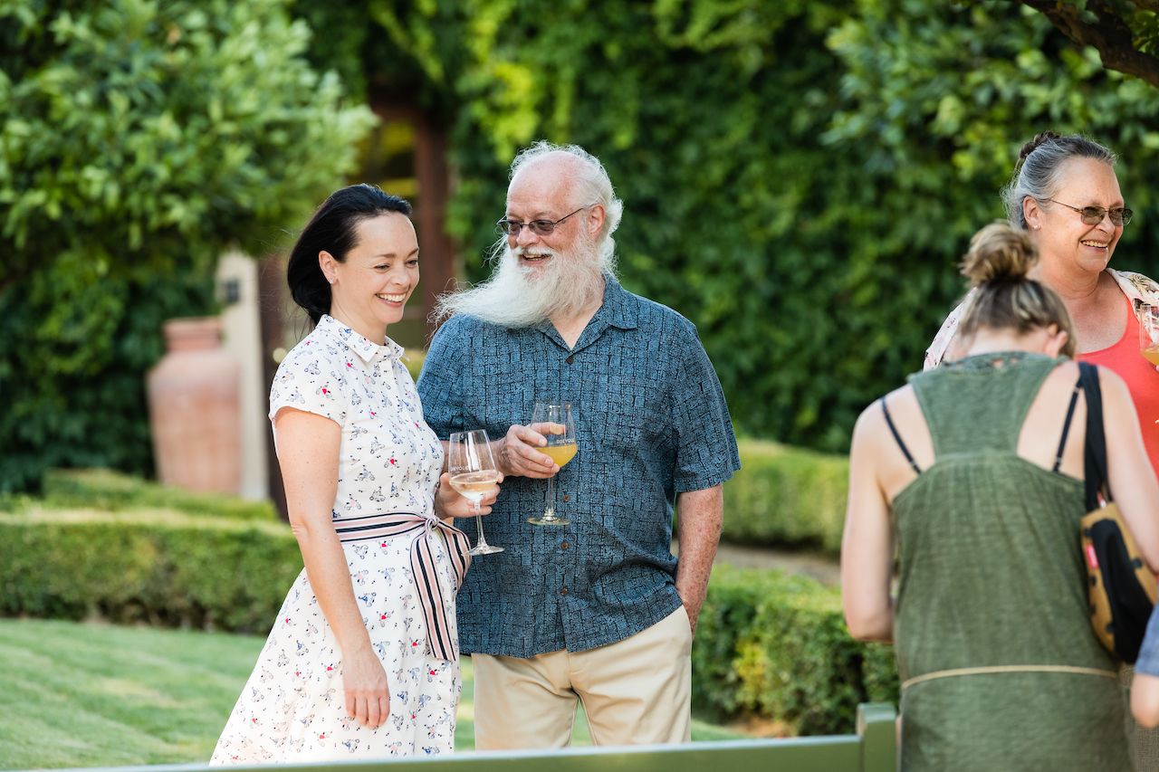 A man and woman smile while holding drinks and chatting at an outdoor wedding welcome barbecue.