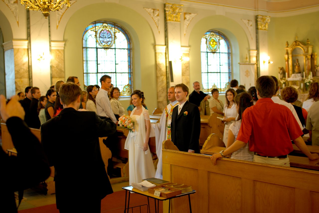 The bride and groom stand at the front of a church, surrounded by guests, as a photographer takes a picture.