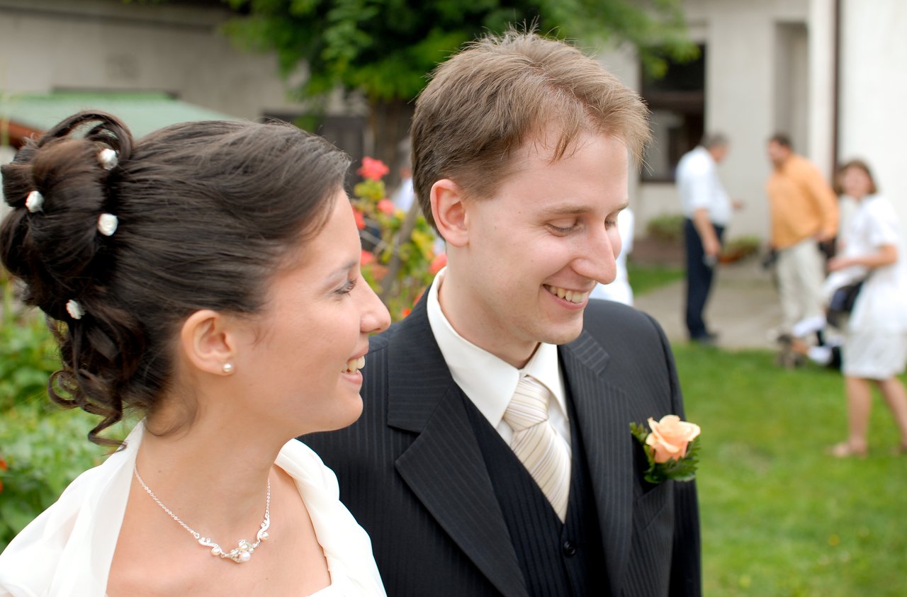 A smiling bride and groom stand together outdoors on their wedding day, dressed in formal attire.
