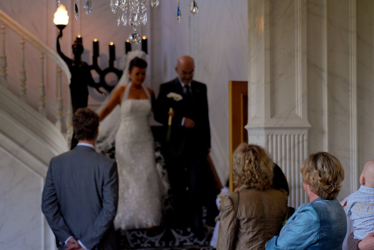 A bride in a white dress walks down the stairs with an older man, as guests watch.
